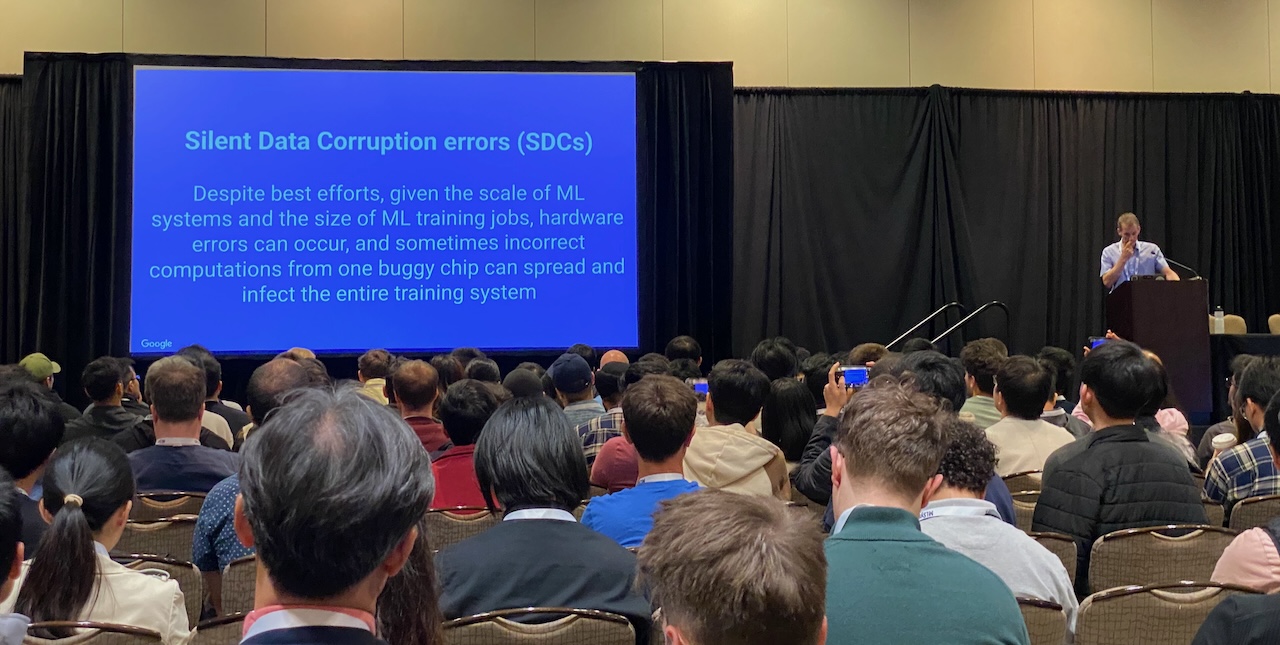Фотография Джеффа Дина с докладом на MLSys 2024. На сцене он стоит у кафедры; на проецируемом слайде с заголовком «Silent Data Corruption errors (SDCs)» объясняется, что аппаратные ошибки в масштабе ML могут распространяться и заражать всю обучающую систему.